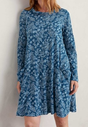 Vrouw draagt een blauw, knielang jurkje met lange mouwen en lichtblauwe bloemen- en bladvormen, staande tegen een effen muur.