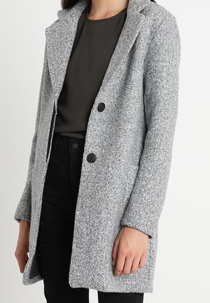Femme portant un manteau en laine gris clair boutonné sur un haut foncé et un pantalon noir, debout devant un fond blanc uni.