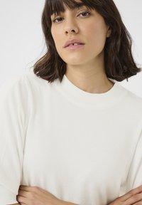 Valge nokk-sömaline topp ümmarguse kaeluse ja pehmete puhvadega lühikeste varrukatega. Sile tekstuur, keha lähedane siluett ja minimalistlik disain.