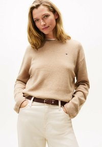 Beige gestrickter Pullover mit langen Ärmeln, rundem Halsausschnitt und einem kleinen Logo. Kombiniert mit hellen Hosen und einem braunen Gürtel.