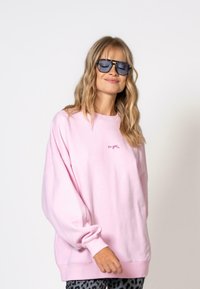 Svetloružová oversized mikina s rebrovanými manžetami a lemom, s nápisom "OH APRIL" v kontrastnej farbe. Model má na sebe tmavé slnečné okuliare.