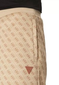 Joggers beige con un patrón repetitivo de logotipos estilizados en forma de diamante, que cuentan con un bolsillo lateral y un parche de logotipo en forma de triángulo rojo.