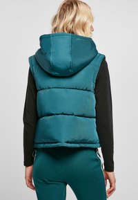 Gilet rembourré teal avec capuche. Présente un design matelassé et des accents de cordon élastique à la base. Porté par-dessus un haut à manches longues noir.