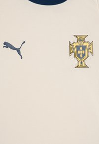Maglia da sport color crema con colletto a girocollo blu navy. Presenta un logo Puma blu scuro e uno stemma d'oro della nazionale portoghese sul lato sinistro.