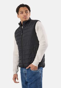 Unge mand iført sort quiltet vest over lys sweatshirt og blå jeans, står med den ene hånd i lommen og ser til siden.