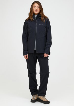 Kvinde iført en sort lynlåstrøje, sorte bukser og beige vandresko, stående mod en ensfarvet lys baggrund.