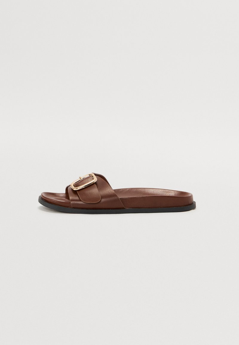 Chinelo de couro castanho com uma faixa larga e um detalhe de fivela dourada, sola plana e textura suave.