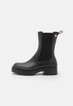 Bottes chelsea noires avec un corps en caoutchouc lisse, des panneaux latéraux élastiques et une semelle épaisse texturée. Dispose d'un tirant pour un enfilage facile.