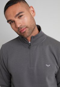 Grauer Reißverschluss-Sweatshirt mit hohem Kragen, geripptem Textil und einem dekorativen weißen Logo auf der Brust. Weicher Stoff, lässiges Design.