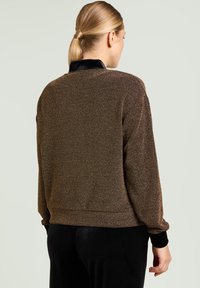 Braune, strukturierte Jacke mit einem schwarzen, samtigen Kragen, langen Ärmeln und einem taillierten Bund, von hinten vor hellem Hintergrund gezeigt.