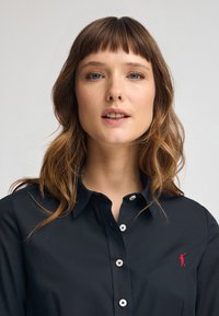 Camicia nera a maniche lunghe con colletto, bottoni bianchi e un piccolo logo rosso ricamato sul petto sinistro. Tessuto dalla texture liscia.