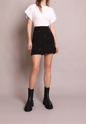 Woman wearing a white short-sleeve top, black mini skirt with small skull patterns, and black chunky ankle boots against a neutral background.