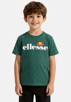 Jeune garçon souriant aux cheveux bruns courts, portant un t-shirt vert avec un logo blanc « ellesse » et un pantalon noir, debout devant un fond uni.