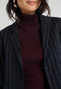 Marinblå randig kavaj med enkel knappdetalj bärs över en vinröd ribbad polo. Kavajen har skarpa slag och strukturerade axlar.