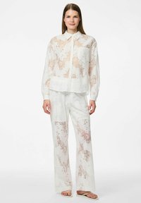 Transparante witte bloemenkant outfit die bestaat uit een blouse met kraag en lange mouwen en brede broekspijpen, versierd met zachte roze patronen.