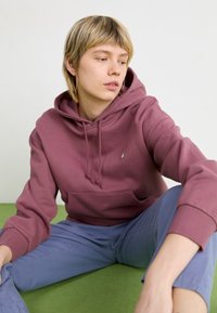 Bordå hoodie med en lommesekk foran, snor og liten logo; brukt med lyseblå bukser på en grønn overflate. Glatt stofftekstur.