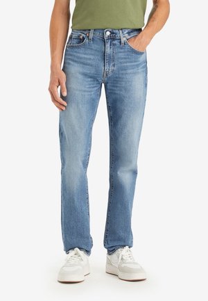 Homme portant un jean droit bleu clair, des baskets blanches et un t-shirt vert olive, une main dans la poche.