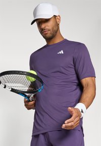 Purple athletic shirt with short sleeves, featuring a white logo. The person holds a tennis racket and a green tennis ball. White cap and wristband accessible.
