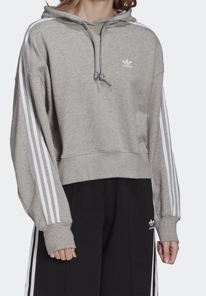 Persoon die een grijze Adidas hoodie draagt met witte strepen op de mouwen en zwarte Adidas broek met witte zijstrepen, staand tegen een effen achtergrond.