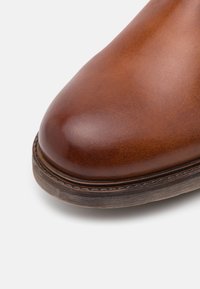 Chaussure en cuir marron à finition lisse, bout arrondi, et semelle texturée contrastante avec des détails de couture visibles.