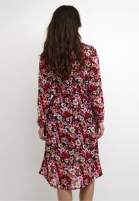 Kukkaisdrawsi kevyessä, puoliksi läpinäkyvässä kankaassa; musta tausta, jossa on kirkkaan vaaleanpunaisia, punaisia ja beigejä kukkia; pitkät hihat ja sidottava vyötärö.