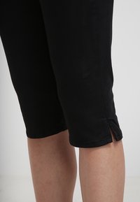 Zbliżenie dolnych nóg w czarnych, krótkich spodniach z rozcięciami po bokach powyżej kolan na jednolitym tle.