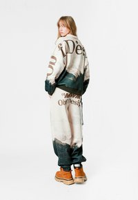 Chaqueta oversized y pantalones a juego con un estampado de paisaje, con acentos en verde oscuro y texto en negrita. Llevado con botas beige.