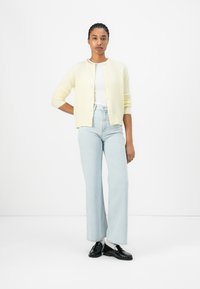 Cárdigan amarillo de Cachemira con cuello redondo y cierre de botones, llevado sobre una blusa blanca y combinado con vaqueros de cintura alta de color azul claro y zapatos negros.