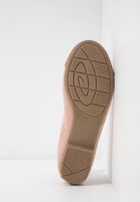 Semelle d'une chaussure plate marron clair présentée en position verticale contre un mur blanc, avec une semelle texturée à motifs géométriques.
