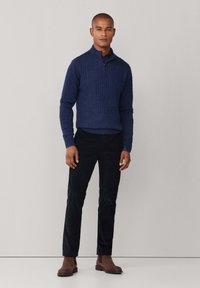 Granatowy sweter w splot włóczkowy z wysokim kołnierzem, w zestawieniu z ciemnymi spodniami slim-fit i brązowymi botkami za kostkę, stojący na neutralnym tle.