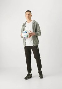 Jonge man in sportkleding met een voetbal, draagt een grijze hoodie, wit T-shirt, zwarte Puma-broek en zwarte voetbalschoenen, staand op een witte achtergrond.