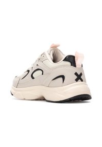 Zapatilla deportiva con parte superior en beige, acentos negros, tira de agarre rosa y suela texturizada. Presenta una forma redondeada y un diseño acolchado.