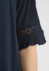 Marinblus med kort ärm som har spetsdetaljer och volanger vid ärmsluten, tillverkad av ett strukturert, lätt tyg.