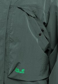 Grüne karierte Jacke aus leichtem Stoff, mit einer Vorderseite-Tasche, zwei schwarzen Knöpfen und einem sichtbaren grünen Logo.