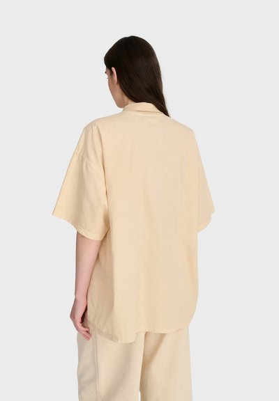 Femme aux longs cheveux foncés, portant une chemise beige oversize à manches courtes et un pantalon large assorti, de dos sur un fond clair uni.