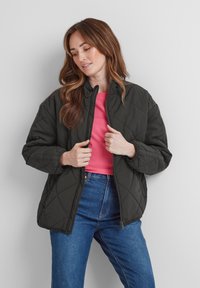 Quilted dark green jacket with a high collar, zip closure, and elastic cuffs, worn over a pink shirt and blue jeans.