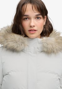 Femme aux cheveux bruns courts portant une doudoune gris clair avec une bordure en fourrure beige autour de la capuche, regardant droit devant elle.