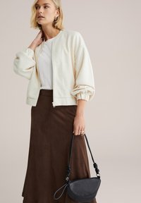 Krémovo sfarbená textúrovaná bomber bunda, biely top, hnedá semišová midi sukňa, drží malú čiernu kabelku s detailmi na skrútenom remienku.