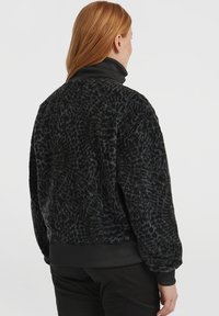 Persoon met rood haar, gekleed in een zwarte, gestructureerde nepbontjas en zwarte broek, staat met de rug naar de camera tegen een effen achtergrond.