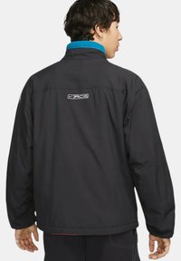 Svart jacka med hög krage, blå fleecefoder och reflektiv logotyp på ryggen. Har en avslappnad passform och lättviktsmaterial.