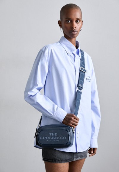 Geantă crossbody din piele albastră marin, cu formă rectangulară, caracteristici de imprimare cu logo, închidere cu fibră și curea ajustabilă. Asortată cu o cămașă cu dungi albastre deschise.
