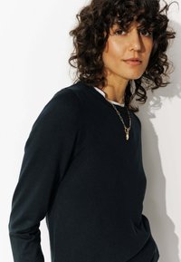 T-shirt en tricot à manches longues bleu marine avec encolure ronde, porté par-dessus une chemise blanche, présentant une texture lisse et une coupe ajustée. Accent avec un collier en or.