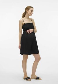 Robe de maternité noire avec des bretelles fines, un corsage texturé, un short ample et un décolleté en scoop, associée à des sandales plates.