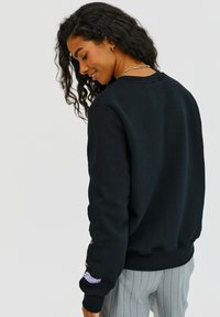 Čierna mikina s rebrovaným okrajom výstrihu a manžetami, zdobená farebnými nášivkami na rukávoch a s jemne česanou textúrou, skombinovaná s pruhovanými nohavicami.