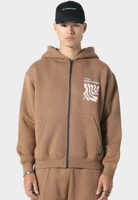 Brauner Reißverschluss-Hoodie aus weichem Material, mit einer Fronttasche, gerippten Bündchen und weißem Logo-Graphic mit Pflanzenmotiv.