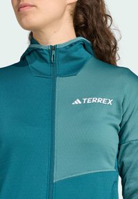 Tyrkysová bunda s kapucňou s hladkou textúrou, zipsom vpredu a kontrastnou svetlejšou tyrkysovou panelou s bielym logom "TERREX".