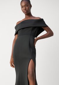 Robe noire dénudée sur les épaules avec une silhouette ajustée, fente latérale, texture de tissu lisse et détail de drapage structuré.