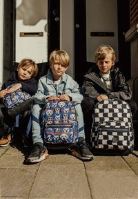 Drie kinderen zitten op de trappen met kleurrijke rugzakken: de ene heeft een patroon van blauwe honden, de andere is zwart-wit geruit.