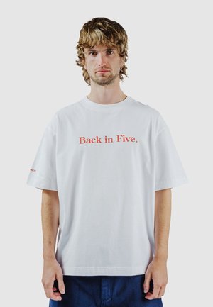 Ung mand med bølget blondt hår iført en hvid, oversize T-shirt med rød tekst "Back in Five." og mørkeblå bukser, stående foran en ensfarvet baggrund.