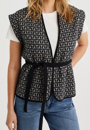 Kvinde iført en sort-hvid mønstret vest med bånd i taljen over en hvid t-shirt og blå jeans, hænderne hviler ved siderne.